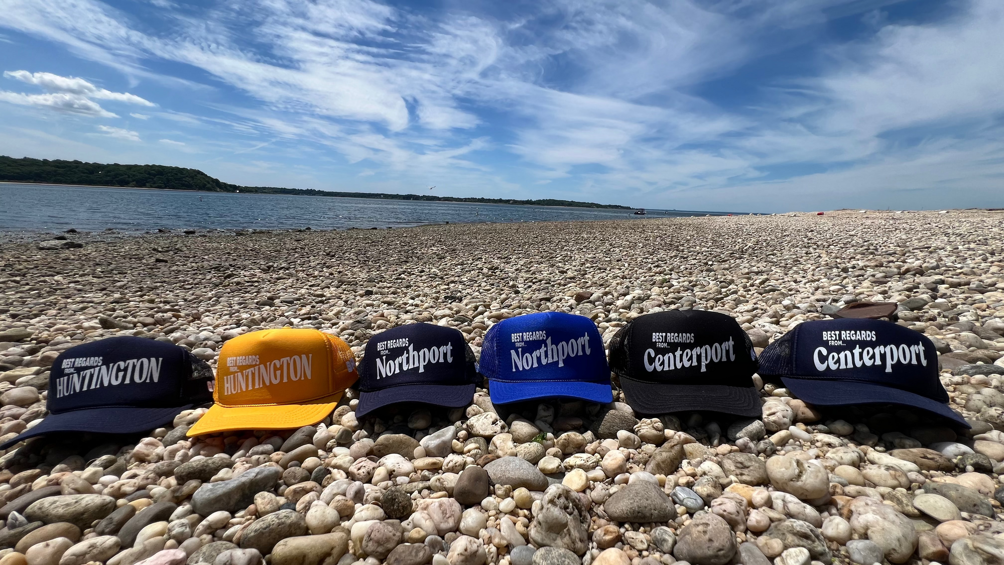 Six trucker hats with 'Northport', 'Centerport', and "Huntington' printed on them are arranged on a pebbly beach with a blue sky.
