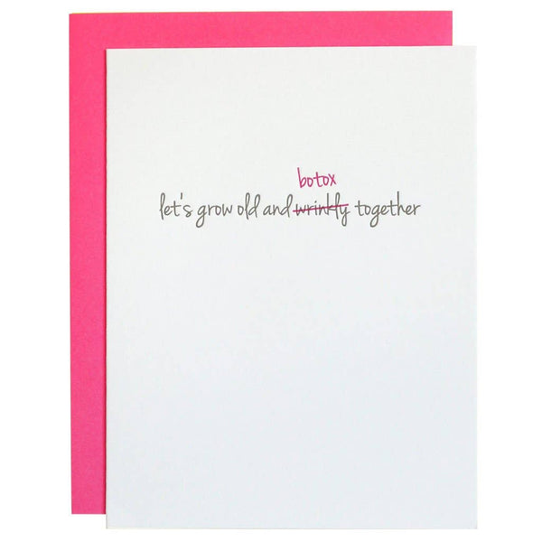 White greeting card with "let's grow old and wrinkly together" in light grey font with wrinkly scratched out in pink and corrected to "botox". Neon pink envelope and blank inside.